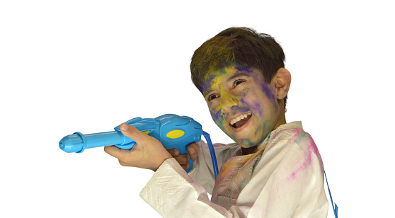 Horizontal photograph of one 10 year old boy in white coloured Kurta with his face covered with messy Holi powder colours, aiming while holding a squirt water gun. Ideal for posters, greeting cards, postcards related to the Indian festival of colors - Holi. There is no text and ample copy space.