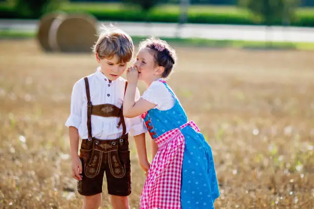 Two kids in traditional Bavarian costumes in wheat field. German children sitting on hay bale during Beer Fest. Boy and girl play at hay bales during summer harvest time in Germany. Best friends