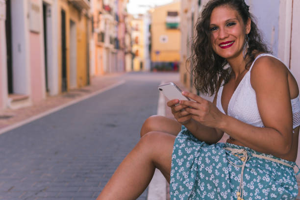 istock A brunette woman sitting smiling at the camera while holding her mobile phone with both hands in the colorful streets of Villajoyosa, Alicante, Spain. 1470448211