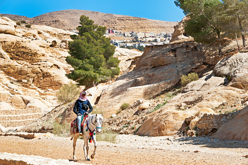 Lone Bedouin rider on horseback in canyon with modern town behind, Petra, Jordan, Middle East. Petra, built around 300 B.C. by the Nabataean Arabs, is the world famous archaeological site in Jordan's southwestern desert. Capital of the Nabatean Kingdom it seamlessly blends Arab style with Hellenistic and Roman or Byzantine architecture. Accessed via Al Siq, a narrow canyon, it contains tombs and temples carved into the pink sandstone cliffs, hence the 'Rose City' The most famous and iconic structure is Al Khazneh, a temple with an ornate Greek-style facade, also known as The Treasury