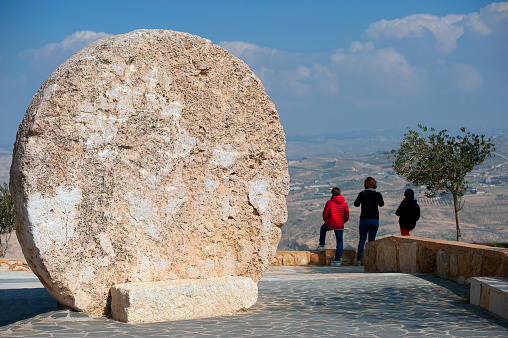 Cave closure stone and tourists enjoying view, Mount Nebo, Abarim mountains, Jordanian Highlands, Jordan, Middle East. This stone is supposed to be similar to the one that closed the cave and was rolled back after Jesus was interred within it. Mount Nebo is an elevated ridge approximately 710 metres above sea level, located in the Jordanian Highlands, a part of the Abarim mountain range, it is mentioned in the Bible Old Testament as the place where Moses was granted a view of the Promised Land before his death. The Church of Moses is situated here and he is reputed to have been buried somewhere on the mountainside with sweeping views of the Holy Land.