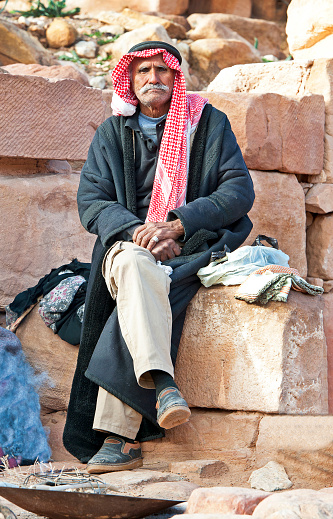 Lone Bedouin figure rests by campfire in Colonnaded Street, Petra, Jordan, Middle East. Petra, built around 300 B.C. by the Nabataean Arabs, is the world famous archaeological site in Jordan's southwestern desert. Capital of the Nabatean Kingdom it seamlessly blends Arab style with Hellenistic and Roman or Byzantine architecture. Accessed via Al Siq, a narrow canyon, it contains tombs and temples carved into the pink sandstone cliffs, hence the 'Rose City' The most famous and iconic structure is Al Khazneh, a temple with an ornate Greek-style facade, also known as The Treasury
