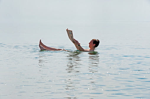 Recreational swimmer floats whilst reading newspaper in the Dead Sea,, Jordan Valley, Jordan, Middle East. The Dead Sea is a salt lake bordered by Jordan to the east and the West Bank and Israel to the west, lying in the Jordan Rift Valley, its main tributary being the Jordan River. The lake's surface is 430.5 metres (1,412 ft) below sea level making its shores the lowest land elevation on Earth anf the deepest hypersaline lake, saltiest bodies of water,  in the world, 10 times as salty as the ocean making swimming similar to floating. This  makes for a harsh environment in which plants and animals cannot survive, hence its name.