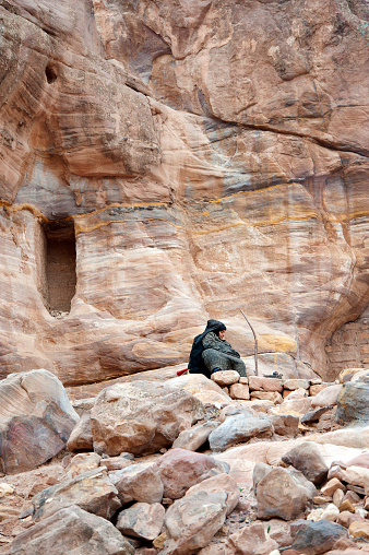 Lone Bedouin woman seated by campfire outside shelter,Colonnaded Street, Petra, Jordan, Middle East. Petra, built around 300 B.C. by the Nabataean Arabs, is the world famous archaeological site in Jordan's southwestern desert. Capital of the Nabatean Kingdom it seamlessly blends Arab style with Hellenistic and Roman or Byzantine architecture. Accessed via Al Siq, a narrow canyon, it contains tombs and temples carved into the pink sandstone cliffs, hence the 'Rose City' The most famous and iconic structure is Al Khazneh, a temple with an ornate Greek-style facade, also known as The Treasury