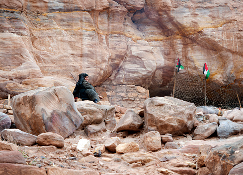 Lone Bedouin figure rests outside her shelter in the Street of Facades, Petra, Jordan, Middle East. Petra, built around 300 B.C. by the Nabataean Arabs, is the world famous archaeological site in Jordan's southwestern desert. Capital of the Nabatean Kingdom it seamlessly blends Arab style with Hellenistic and Roman or Byzantine architecture. Accessed via Al Siq, a narrow canyon, it contains tombs and temples carved into the pink sandstone cliffs, hence the 'Rose City' The most famous and iconic structure is Al Khazneh, a temple with an ornate Greek-style facade, also known as The Treasury