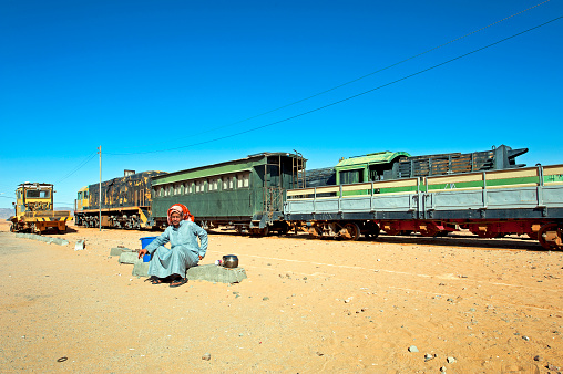 Trader awaits customers for tea at old rail yard, Wadi Rum, Southern Jordan, Jordan, Middle East. This rolling stock was used in the film Lawrence of Arabia and then left to rust. Wadi Rum is a desert valley cut into the sandstone and granite cliffs in southern Jordan. Known also as the Valley of the Moon it has been inhabited since prehistoric times (witnessed by the petroglyph inscriptions) and by semi nomadic tribes since the Nabataeans but is best known for its association with T E Lawrence - Lawrence of Arabia - who based himself here to encourage the Arab Revolt and to attack Turkish Forces of the Ottoman Empire during WW1. He was later immortalized by the film of the same name, made in 1962 by David Lean of which this image shows rolling stock used in the film and later abandoned. Today the only inhabitants are Bedouin tribes and transient tourists.