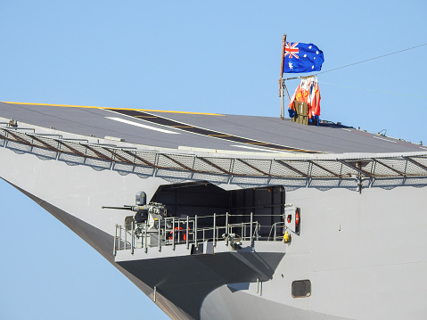The flight deck curve of HMAS Canberra, one of two Amphibious Assault Ships of the Royal Australian Navy.  She is docked at Garden Island, Sydney Harbour.   The colourful objects under to the Australian flag on the flight deck of HMAS Canberra are ship's flags to be deployed over the ship for the public open day the next day, as part of Navy Week.  The crown on top of the flagpole means the flotilla commander is based on this ship.  One of her six 12.7mm machine guns has the barrel uncovered and pointing out in the direction of the Sydney Harbour Bridge.  This image was taken on a sunny afternoon from Woolloomooloo Bay on 25 February 2023.