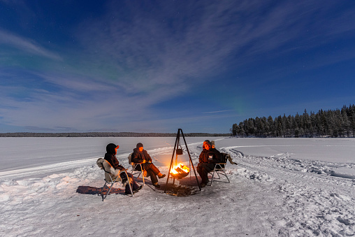 View of a family of three people sitting around the campfire above a frozen lake under northern lights