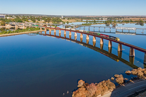 Murray Bridge, Australia - Feb 20, 2023: Aerial view colourful Pacific National interstate container freight train with 2 diesel locomotives travelling from Perth to Melbourne via Adelaide. The train is reflecting in the mirror-calm floodwater alongside the River Murray at Murray Bridge as it crosses the viaduct in South Australia. The swollen river breached a levee on January 5, rapidly filling the agricultural farming land with levels of water not seen since the great flood of 1956. The second locomotive is painted in the distinctive red 