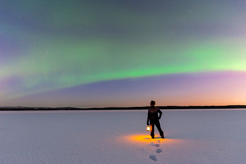 Woman with lamp in her hand watching northern lights on a frozen and snowy lake under a clear sky, Swedish Lapland, Sweden