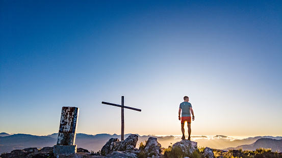 One male trail runner hiker silhouette looking sideways standing on a rock next to a cross and cairn mountain beacon on a mountain summit at sunrise, Simonsberg, Stellenbosch, Cape Winelands, Western Cape, South Africa.