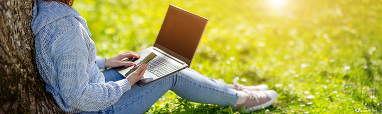 Woman with laptop sitting on the grass in the natural park. Concept of the freelance, working and learning.