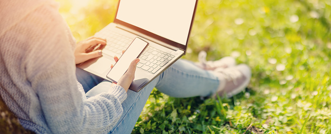 Woman with laptop and smartphone sitting on the grass in the natural park. Concept of the freelance, working and learning.