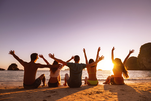 Group of happy young friends are having fun and sits on the sand at sunset beach