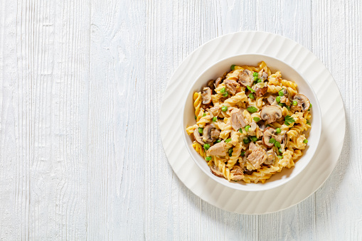 tuna mushroom green pea fusilli bake with creamy cheese sauce in white bowl on white wood table, flat lay, free space