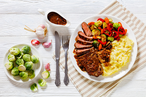 medium rare beef steak with brown gravy and mashed potato, roasted brussels sprouts, pepper and carrots on white plate on wooden table with cutlery, horizontal view from above, flat lay