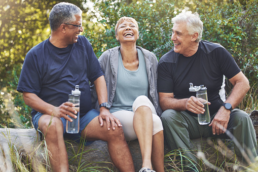 Nature, fitness and senior friends in conversation while sitting in the forest after hiking. Happiness, communication and elderly people talking, bonding and drinking water after outdoor exercise.
