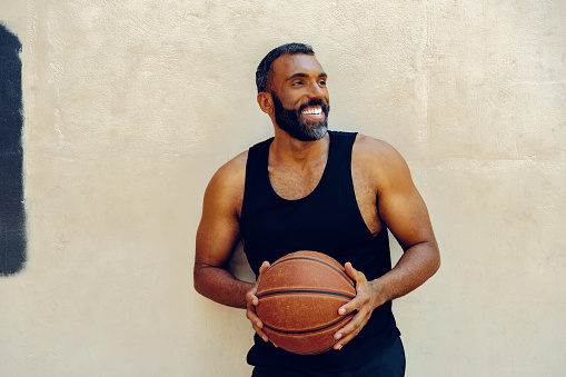 A bearded male athlete exercises indoors, dribbling a basketball. He wears a sleeveless tank top and looks determined in his pursuit of success.