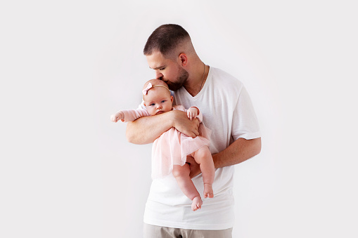 Portrait father holding his newborn baby girl on white background. Dad kissing small daughter in pink dress