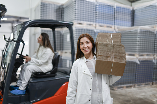 Smiling Female Warehouse Workers Carrying Cardboards And Driving Forklift