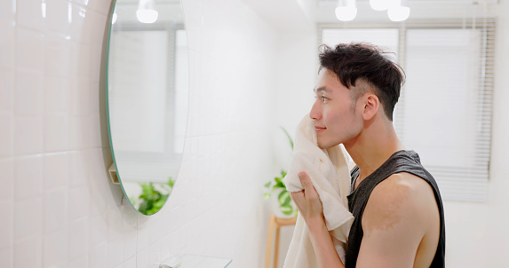 side view asian cheerful man standing in front of mirror is wiping his face with towel after washing face