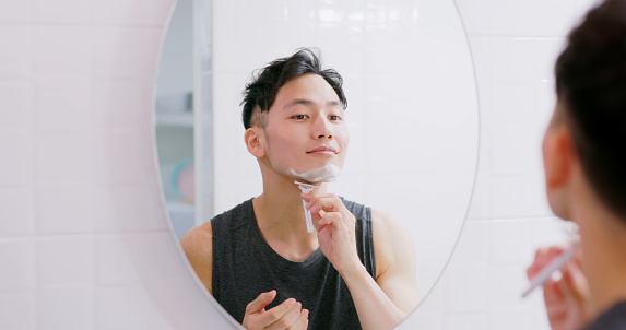 asian man standing in front of mirror is using razor and shaving face with foam