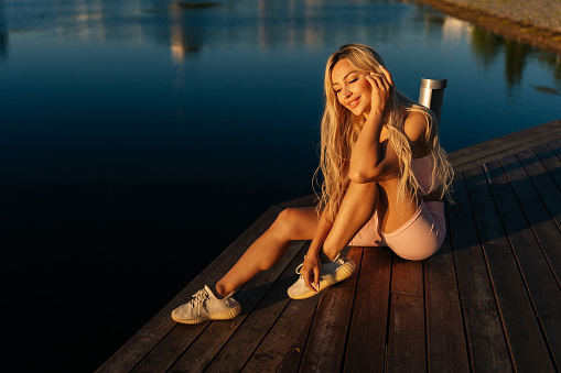High-angle view of cheerful sporty blonde woman in sportswear sitting posing on wooden pier cite park by water on sunny summer morning, looking down. Concept of outdoor sport and healthy lifestyle.