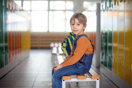 Sweet blonde toddler boy standing in front of a lockers in kindergarden or school hallway, wintertime