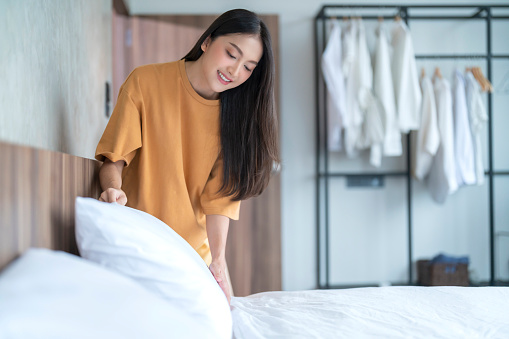 Happy Asian young adult female woman setup her bed sheet blanket by herself in the morning with sunlights from window at home apartment