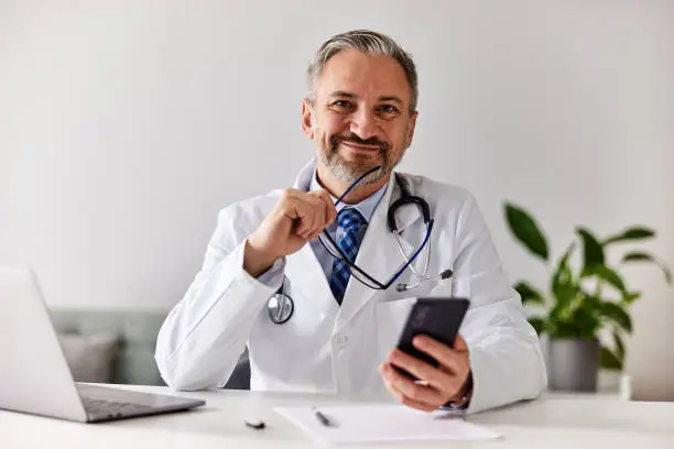 A smiling senior male doctor sitting in front of the laptop, holding a smartphone and gasses, looking at the camera.