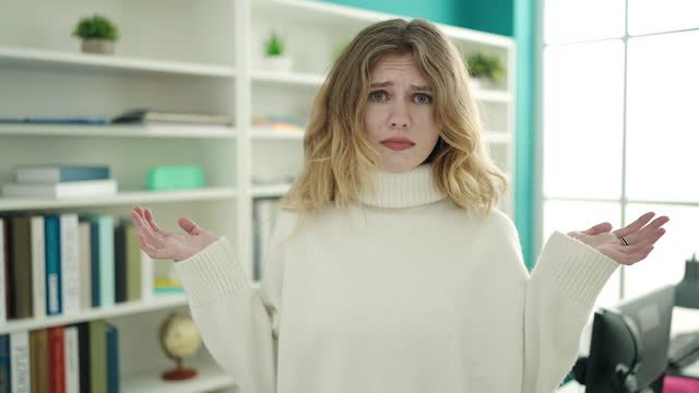 Young blonde woman student standing with confused expression at library university