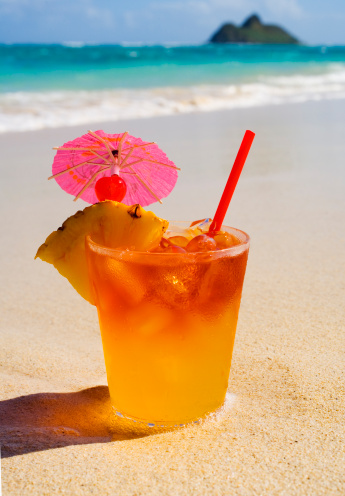 a tropical Maitai cocktail from Hawaii with a garnish of pineapple and a paper umbrella sits on the sands of a Hawaii beach near water's edge