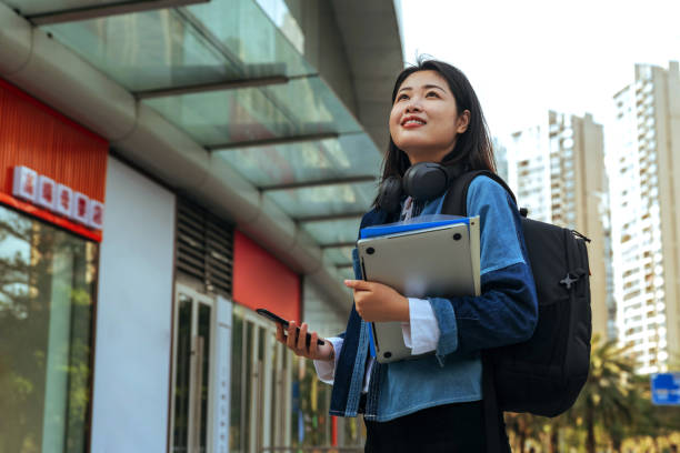 cute female college student looking up while standing outdoors during the day. - looking up young women outdoors laptop imagens e fotografias de stock