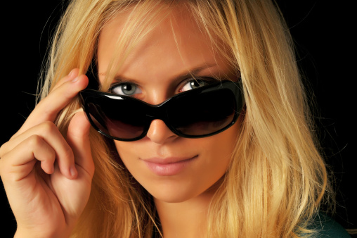 Portrait of beautiful blonde woman peering over dark sunglasses on black background.