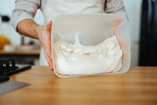 A cropped photo of an unrecognizable man holding a plastic box with dough and turning it over on the worktop.