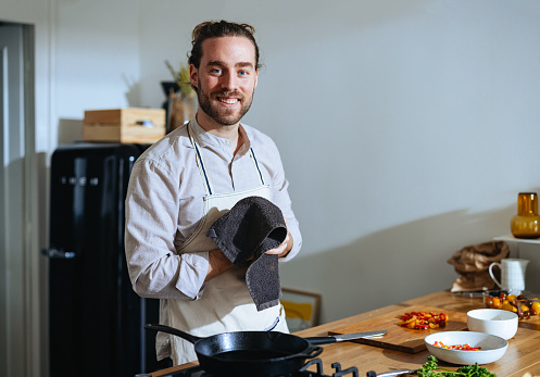 Portrait of a handsome man standing in his kitchen in the morning and preparing a meal. He is looking at the camera, smiling and wiping his hands on a kitchen towel.