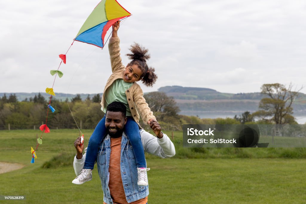Kite Flying is So Much Fun A mid adult father walking outdoors wearing casual clothing with his young daughter sitting on his shoulders, she is flying a multi-coloured kite in the air, they are smiling and having fun together while in Scotland. Kite - Toy Stock Photo