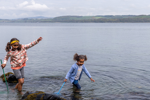 A shot of two young sisters wearing casual clothing stepping in a lake with fishing nets, they are searching the lake to discover something interesting in Scotland.