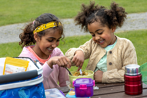 A front-view close-up shot of two young girls sitting at a table outdoors wearing casual clothing while eating their lunch, they are sharing a tub of pasta, they are sisters, smiling and having fun while in Scotland.