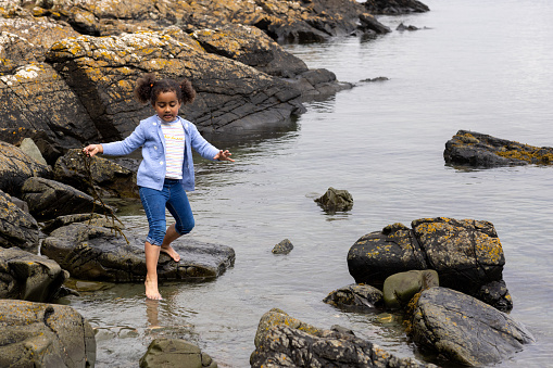 A shot of one young girl wearing casual clothing stepping in a lake, she is holding seaweed in her hand as she tries not to slip on the rocks in Scotland.