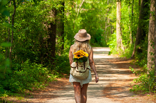 A young woman wearing a hat walks through a trail in the forest with sunflowers in her backpack during a sunny summer day