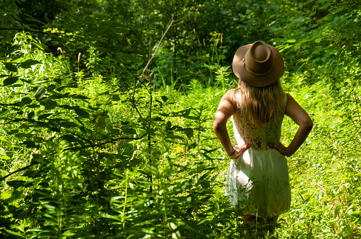 A young woman in a white dress wearing a hat has her arms on her hip in a meadow in the forest on a sunny day