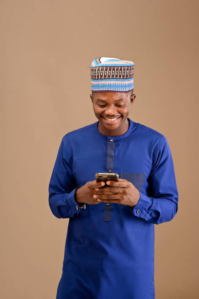istock A smart looking young man smiling and using his smartphone with his two hands, African man wearing blue attires and a cap on isolated brown background 1470540221