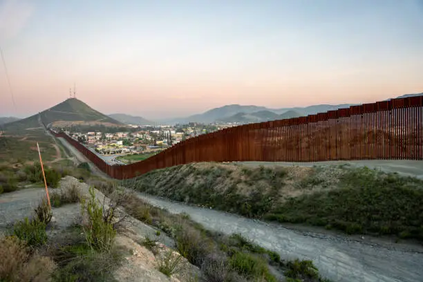 Photo of International Border Wall Between Tecate California and Tecate Mexico Near Tijuana Baja California Norte at Dusk Under Stunning Sunset with View of the City From the USA