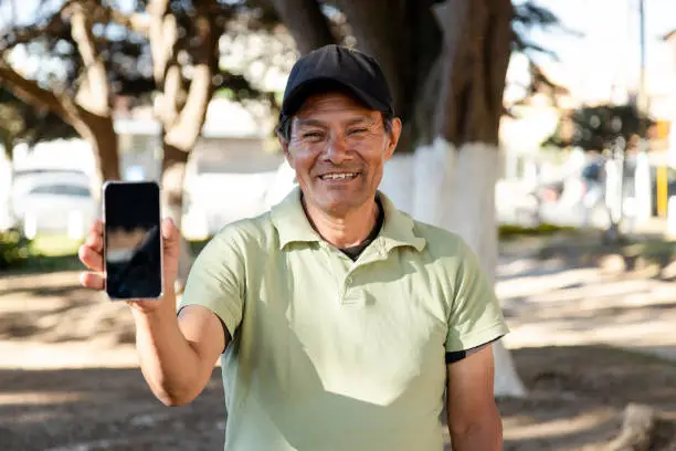 Photo of Hispanic man smiling with phone in hand - Senior adult showing a phone in the park