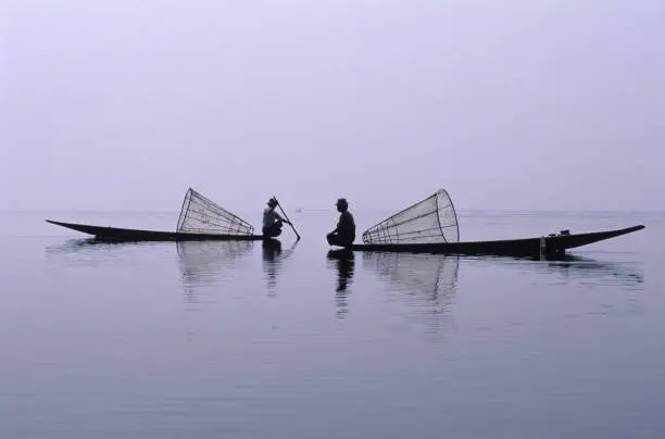 Two Intha leg-rower fishermen on Inle Lake, Myanmar (Burma).  The men sit in traditional flat-bottomed boats facing each other. Behind each man is a typical cone-shaped fish trap. The fishermen and boats are silhouetted against the pale mauve monochrome sky and reflected in the lake. Dusk or dawn horizontal color image with copy space.