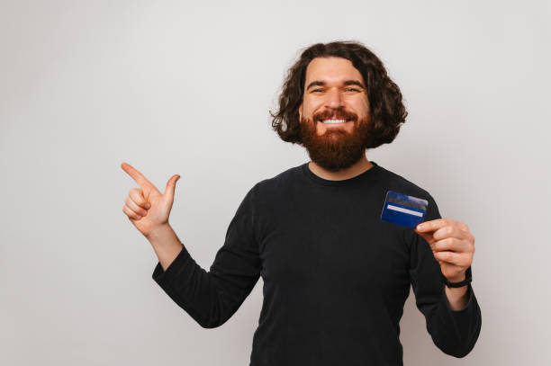 istock Cheerful bearded man is pointing aside and holding a blue credit card. 1470569706