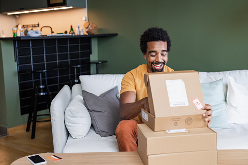 A smiling young man opening parcels at home. He is sitting on the couch in the living room. Cardboard boxes are on the table.