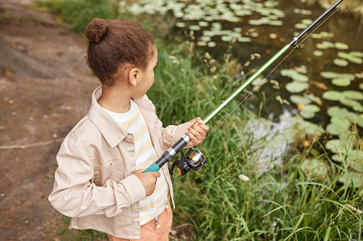 High angle portrait of black little girl fishing by forest lake and enjoying nature, copy space