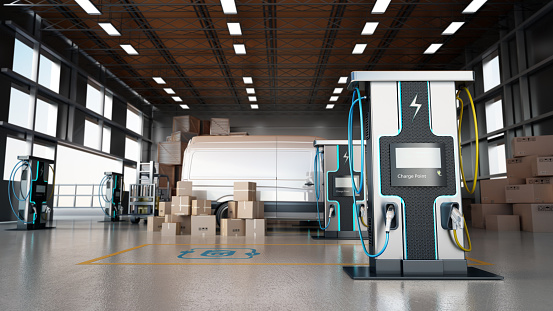 Electric commercial vehicles, industrial charging stations and stack of cargo boxes inside a distribution warehouse.